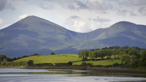 A view across water to the Mourne Mountains in the distance surrounded by foothills and trees